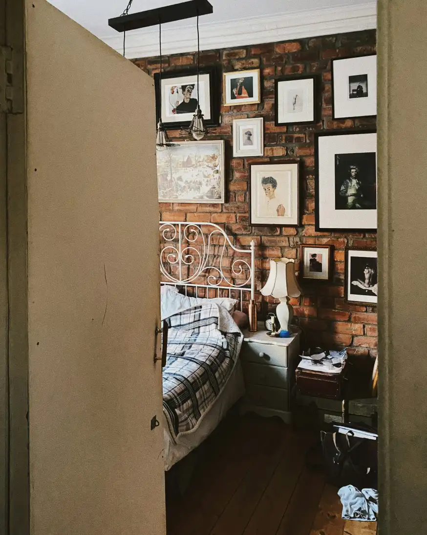 Nhìn qua khung cửa vào phòng Gallery King: tường gạch treo kín tranh và ảnh đóng khung, giường sắt trắng, đèn ngủ