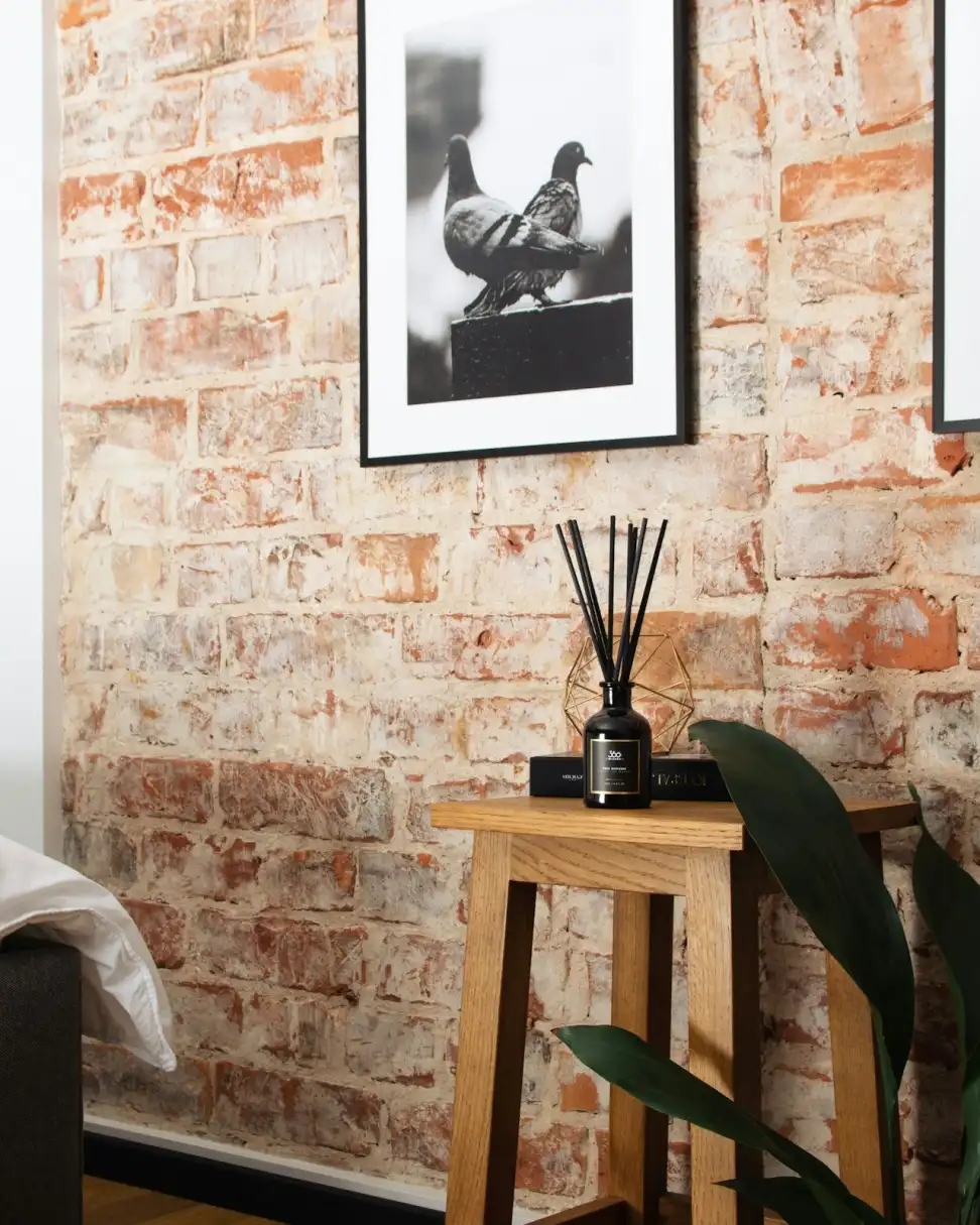 Góc phòng Studio Xưởng: tường gạch thô, ảnh đen trắng đôi chim bồ câu đóng khung, ghế đẩu gỗ và lọ tinh dầu